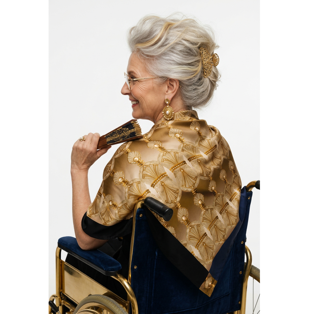 A senior woman with short gray hair and glasses, seated in a wheelchair and shown from the back, wearing a navy blue top and a large gold and beige patterned shawl draped over her shoulders against a plain white studio background.