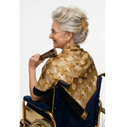A senior woman with short gray hair and glasses, seated in a wheelchair and shown from the back, wearing a navy blue top and a large gold and beige patterned shawl draped over her shoulders against a plain white studio background.