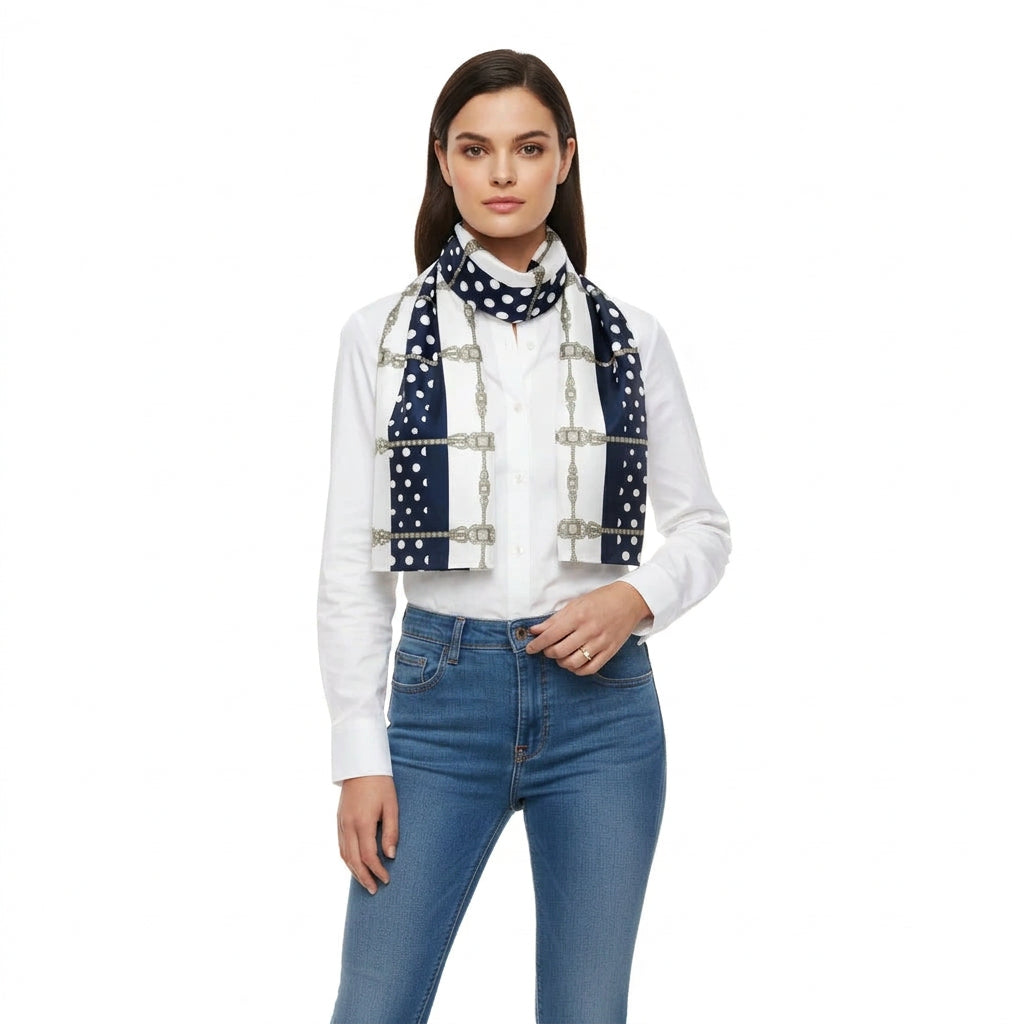 A woman modeling a navy blue scarf with white polka dots and silver-tone chain and buckle details, worn over a white button-down shirt and blue jeans against a plain white background.