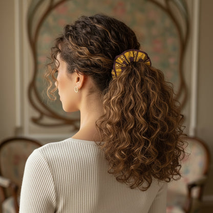 A model with curly hair elegantly tied into a ponytail with a scrunchie featuring a gold Baroque frame pattern on brown recycled fabric.